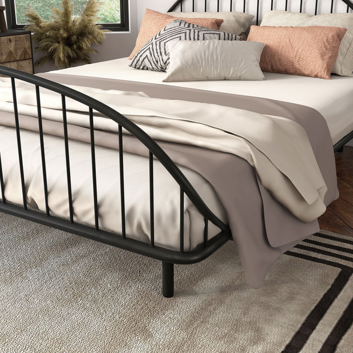 Left facing close up view of an industrial black metal queen bed frame with rounded head and footboards in a room with linens and accessories