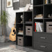 Left facing durable modern three-shelf slim black storage bookcase in a larger modular configuration in a room with accessories