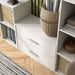 Left facing high-camera view of a modern two-drawer white storage cabinet with silver handles in a larger modular bookcase with accessories