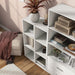 Left facing high-camera view of a durable modern three-shelf slim white storage bookcase in a larger modular configuration in a room with accessories